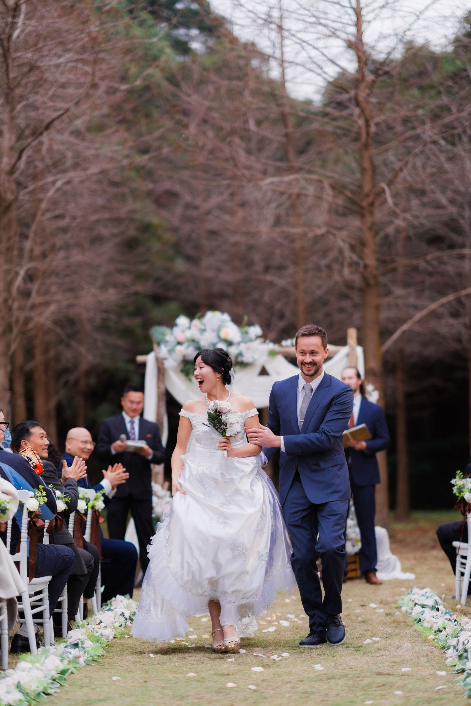 A wedding ceremony processional takes place on an outdoor aisle lined with white flowers and chairs among bare winter trees.