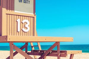 Wooden brown lifeguard stand with 'On Duty' sign stands against bright blue sky on sandy beach.