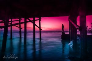 Moody sunset silhouettes a pier over water with vibrant pink and purple skies reflected in the dark waves below.