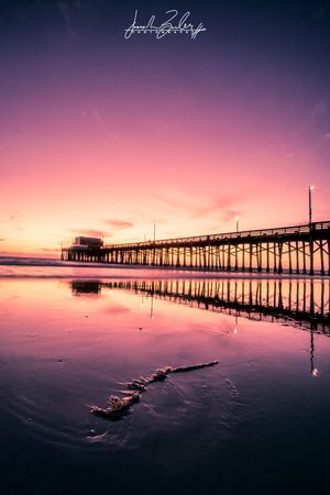 A wooden pier stretches into the ocean at sunset with vibrant pink and purple skies reflecting on calm waters.