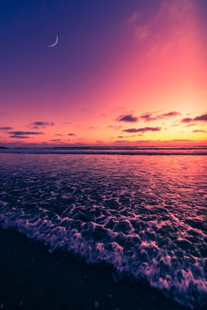 Crashing ocean waves reflect vibrant purple and orange sunset colors with a crescent moon in the dramatic sky.