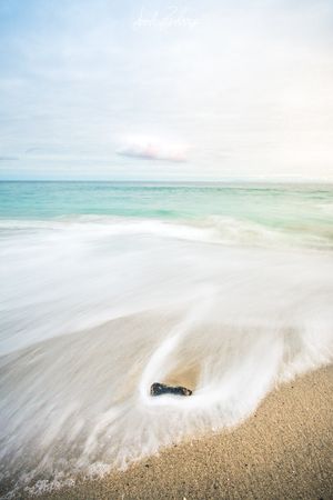 Long exposure photograph of ocean waves washing over a rock on sandy beach with turquoise waters and cloudy sky.