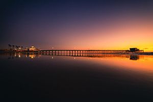 Silhouettes of ocean pier and boats against vibrant orange and purple sunset sky reflecting on calm water.