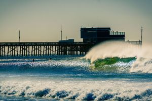 Large waves crash against a pier during sunset with golden light reflecting off the ocean spray.