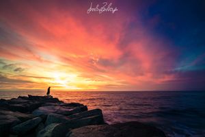 A silhouetted figure stands on rocky jetty against dramatic orange and purple sunset sky over ocean horizon.