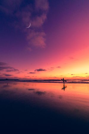 A silhouetted surfer walks on a reflective beach at sunset with a crescent moon in the purple-pink sky.