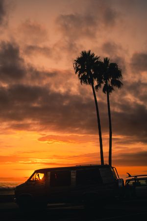 Silhouetted palm trees and vintage van against vibrant orange sunset sky with moody clouds.