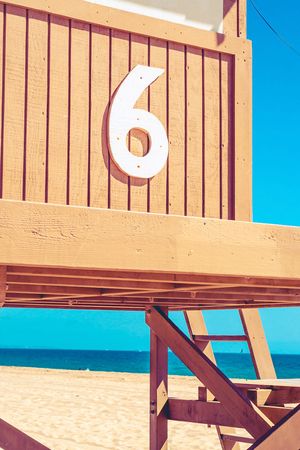 A wooden lifeguard tower with the number 6 in white against tan wood panels and bright blue sea in the background.