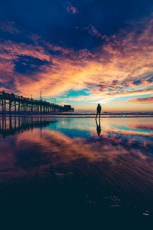 A lone figure stands on a mirror-like beach at sunset near a wooden pier under dramatic orange clouds.