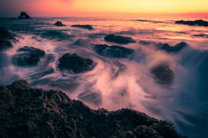 Long exposure seascape capturing misty waves crashing over dark rocks at sunset with vibrant pink sky.