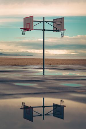 Basketball hoops reflect on flooded court surface against turquoise sky during sunset creating surreal mirror imagery.