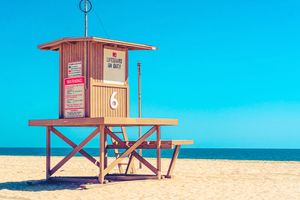 Red and white wooden lifeguard tower stands on sandy beach against bright turquoise sky.