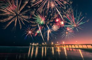 Colorful fireworks display reflecting over calm water at night with a bridge silhouette.