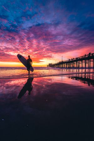 Beach pier with dramatic purple and red clouds at sunset creating a stunning reflection on the wet sand.