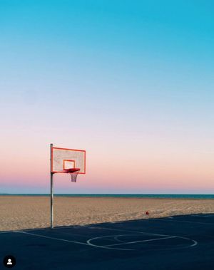 Basketball court and hoop silhouetted against pastel sunset sky on beach.