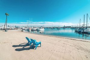 Pristine white sandy beach with blue lounge chairs and boats docked in the marina under clear skies.