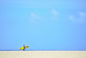 A person wearing yellow carries a surfboard across a white sandy beach against a bright blue sky.