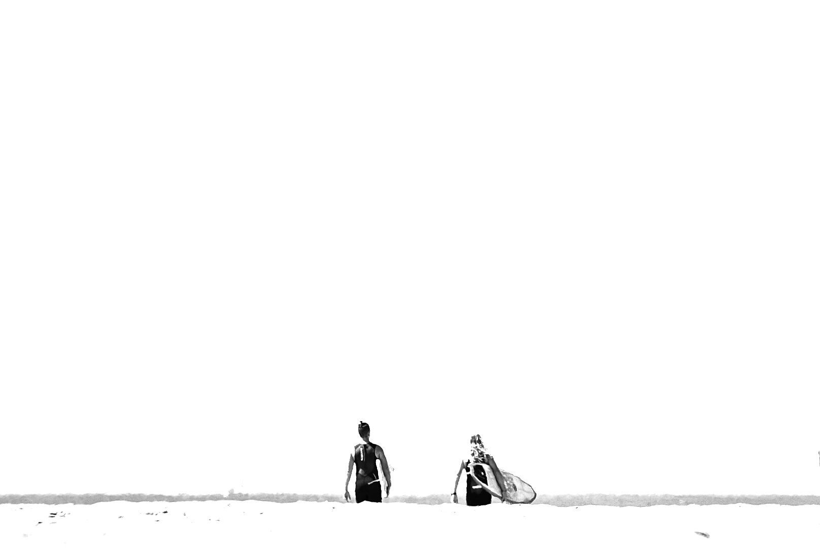 Two surfers walk along an empty beach horizon in minimalist black and white composition.