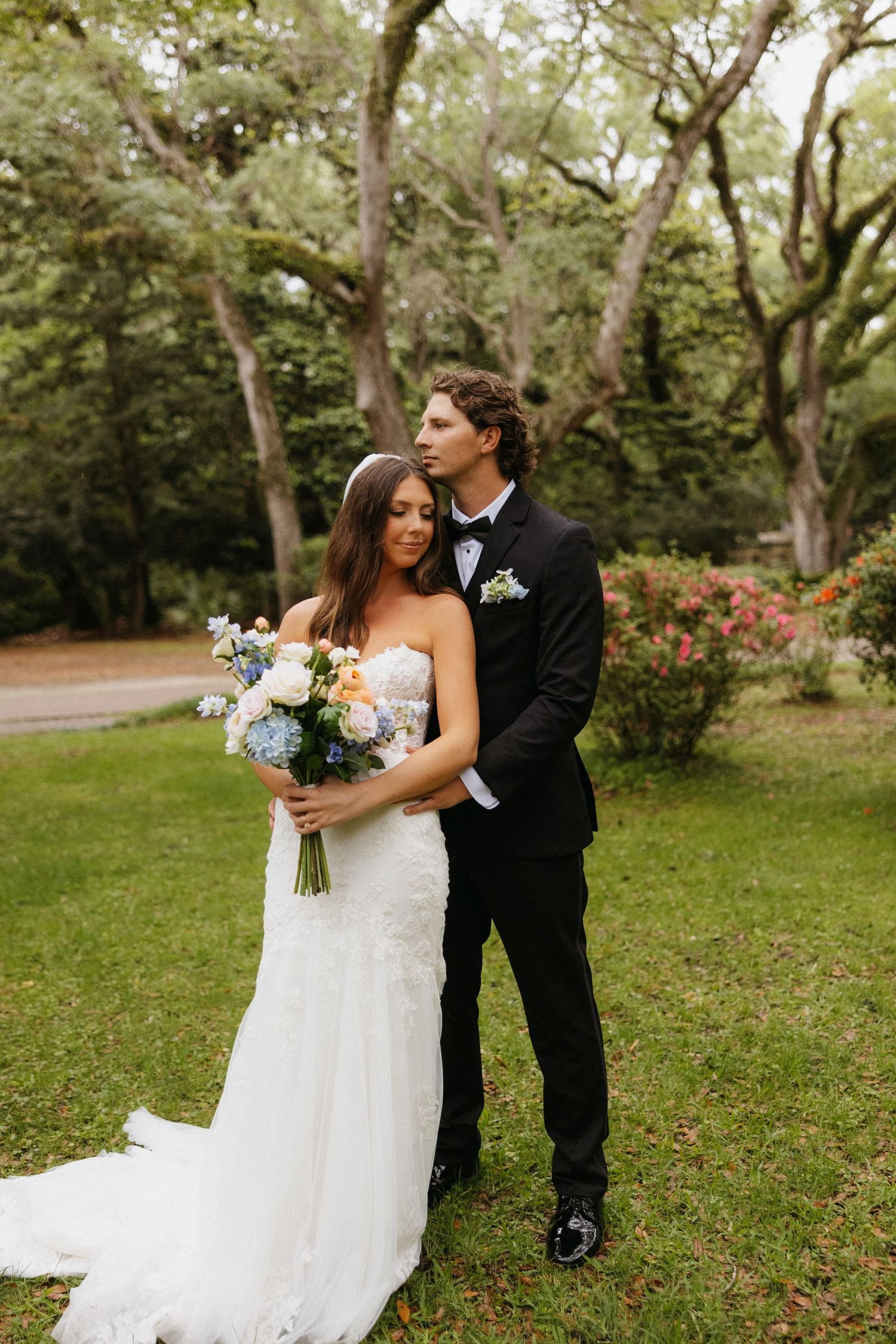 A romantic wedding couple poses outdoors in a lush garden setting with springtime flowers and greenery.