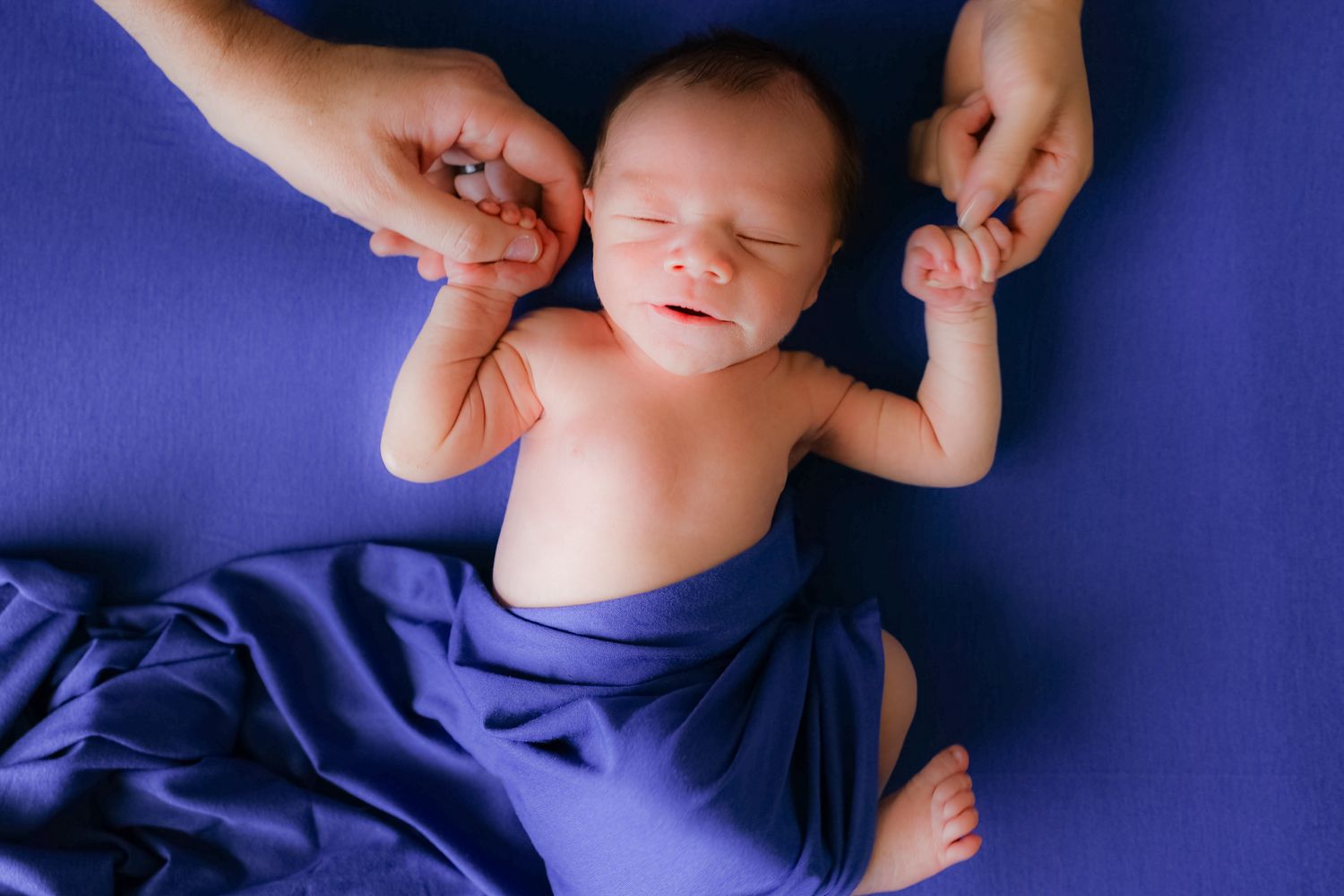 A sleeping newborn baby wrapped in blue fabric being gently held by adult hands.