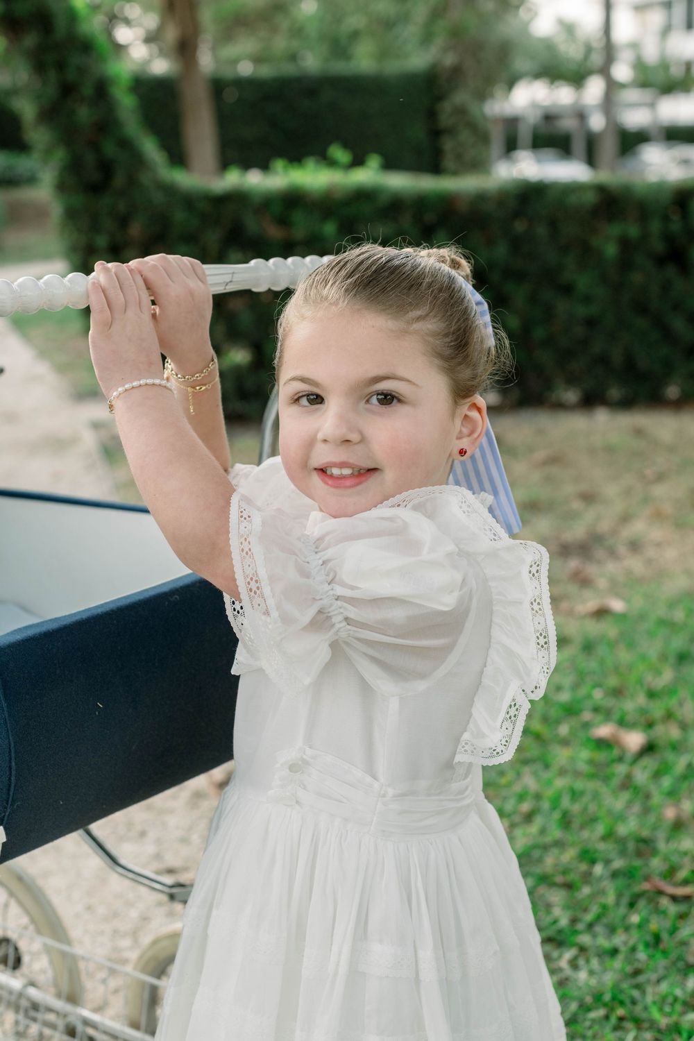 Child in white dress playing with vintage pram in garden setting.