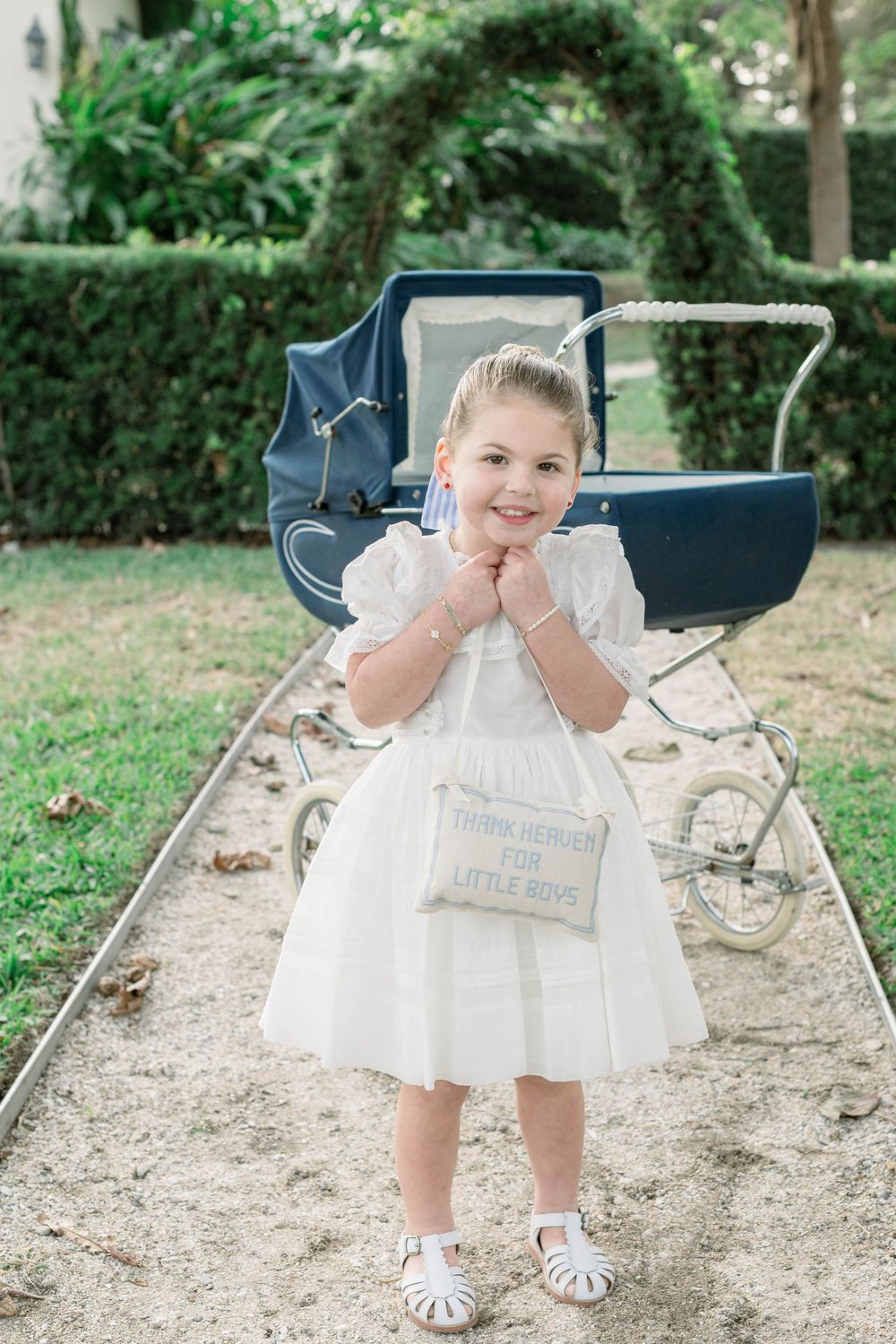 Child in white dress playing near vintage navy pram in garden setting.