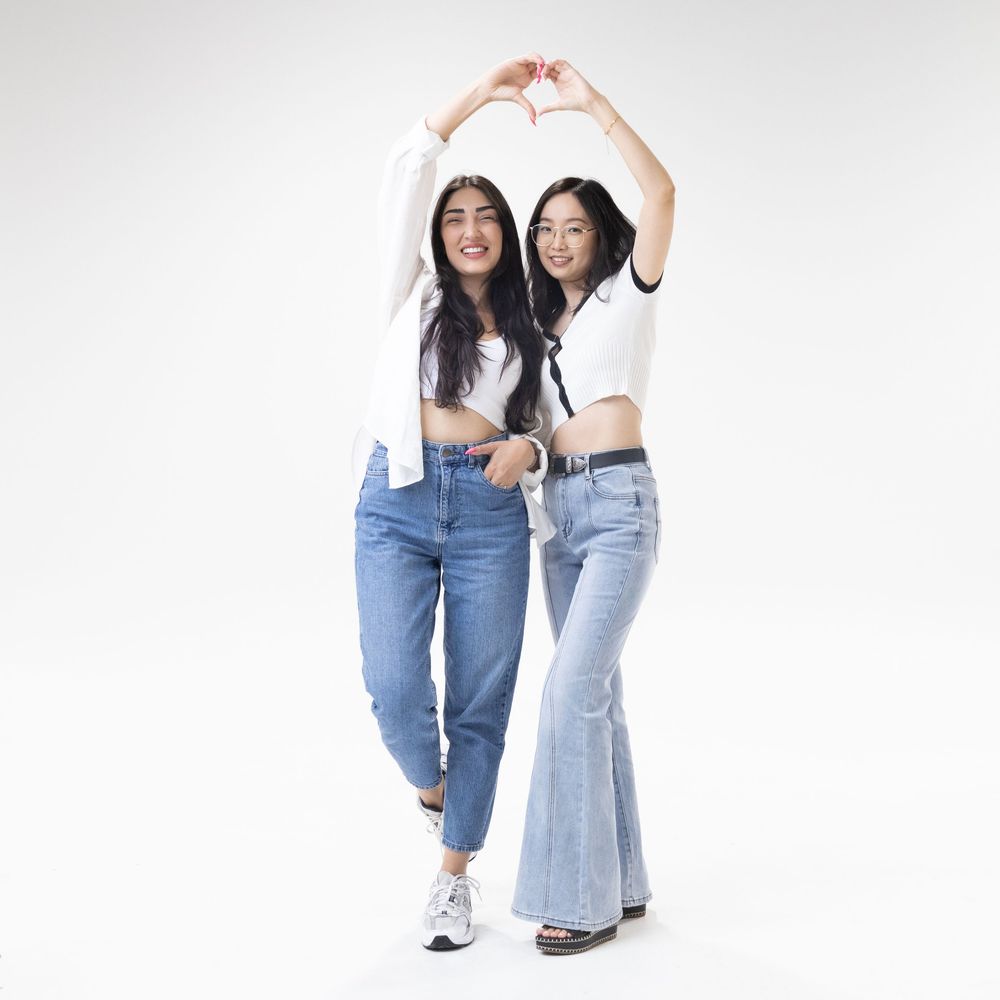 Two women in jeans create heart shapes with their hands while posing together for photos.