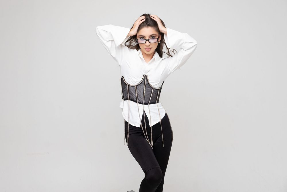 Professional wearing glasses, white button-down shirt with black vest and pants poses with hands behind head on gray background.