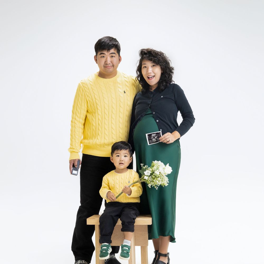A family of three wearing coordinated yellow and green outfits poses together against a white background with flowers.