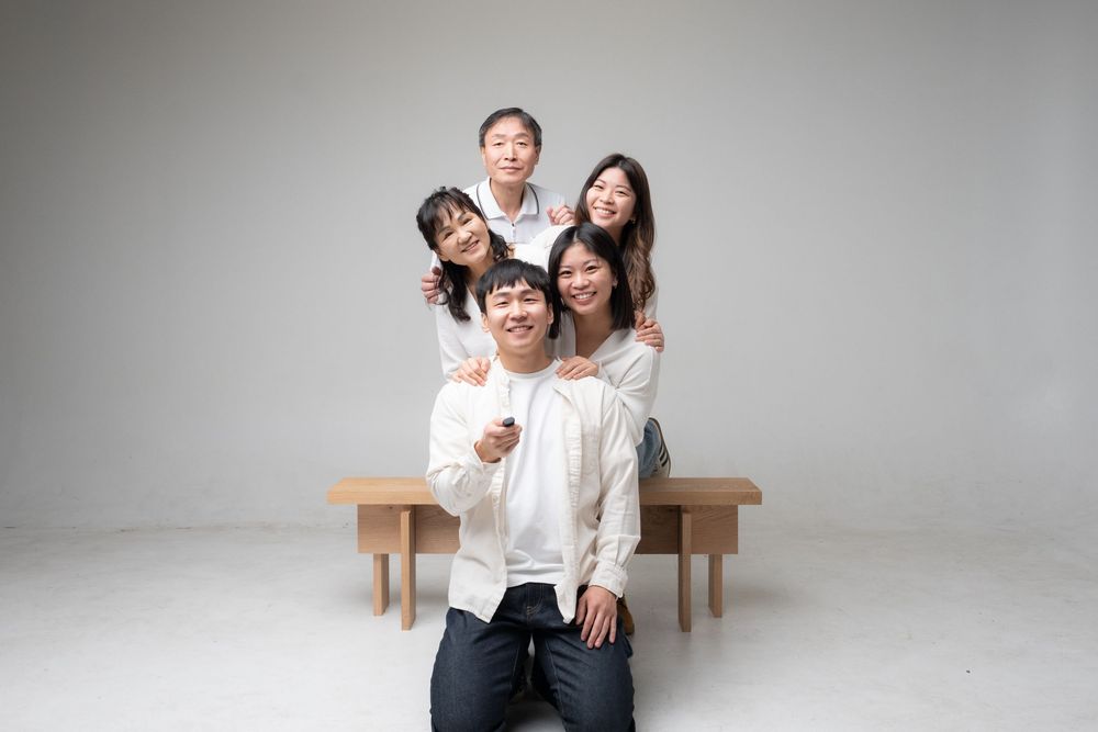 Two people in white shirts and jeans sitting on wooden bench against a light gray background in studio setting.