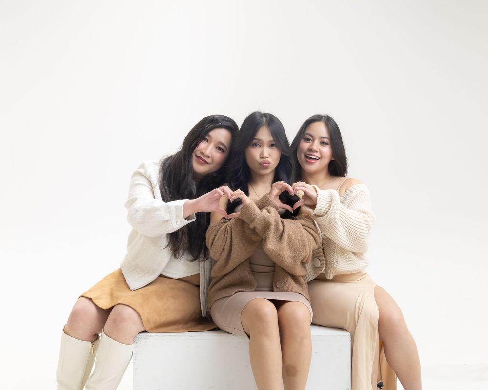 Three friends in cozy sweaters sit together making heart shapes with their hands against a white backdrop.