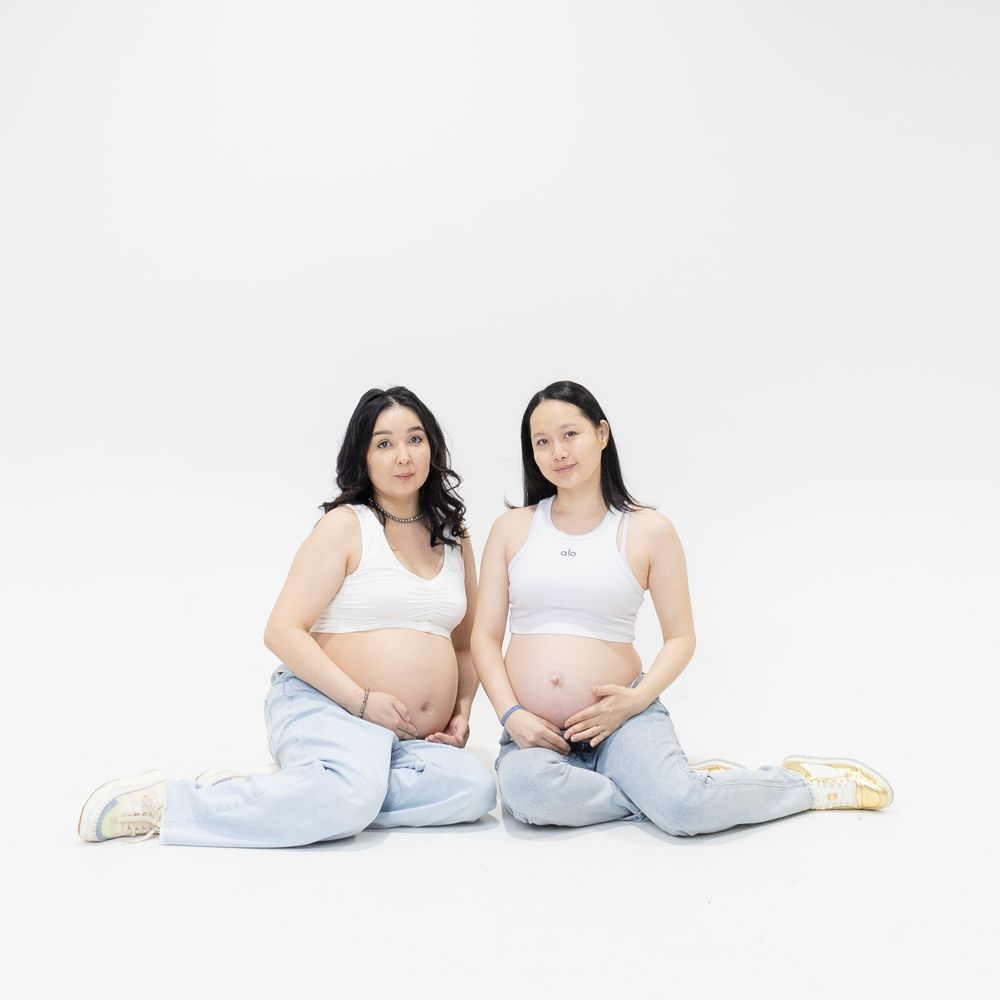 Two pregnant women in white tops and jeans sit cross-legged on a white background.