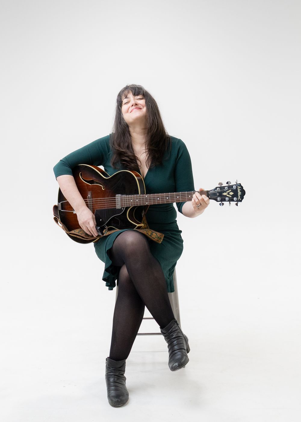Musician in black tights and green top plays acoustic guitar while seated against white studio backdrop in four poses.
