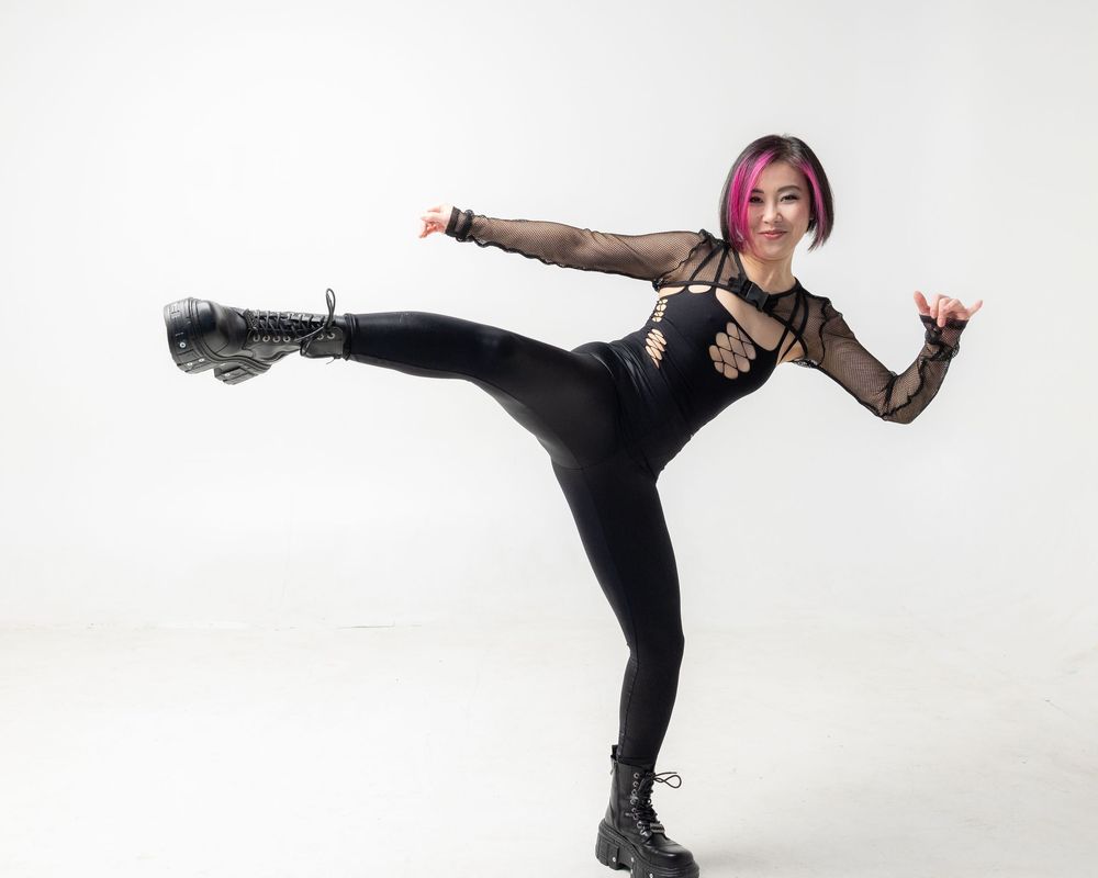 A dancer in black leggings and boots performs a high kick pose against a white studio background.