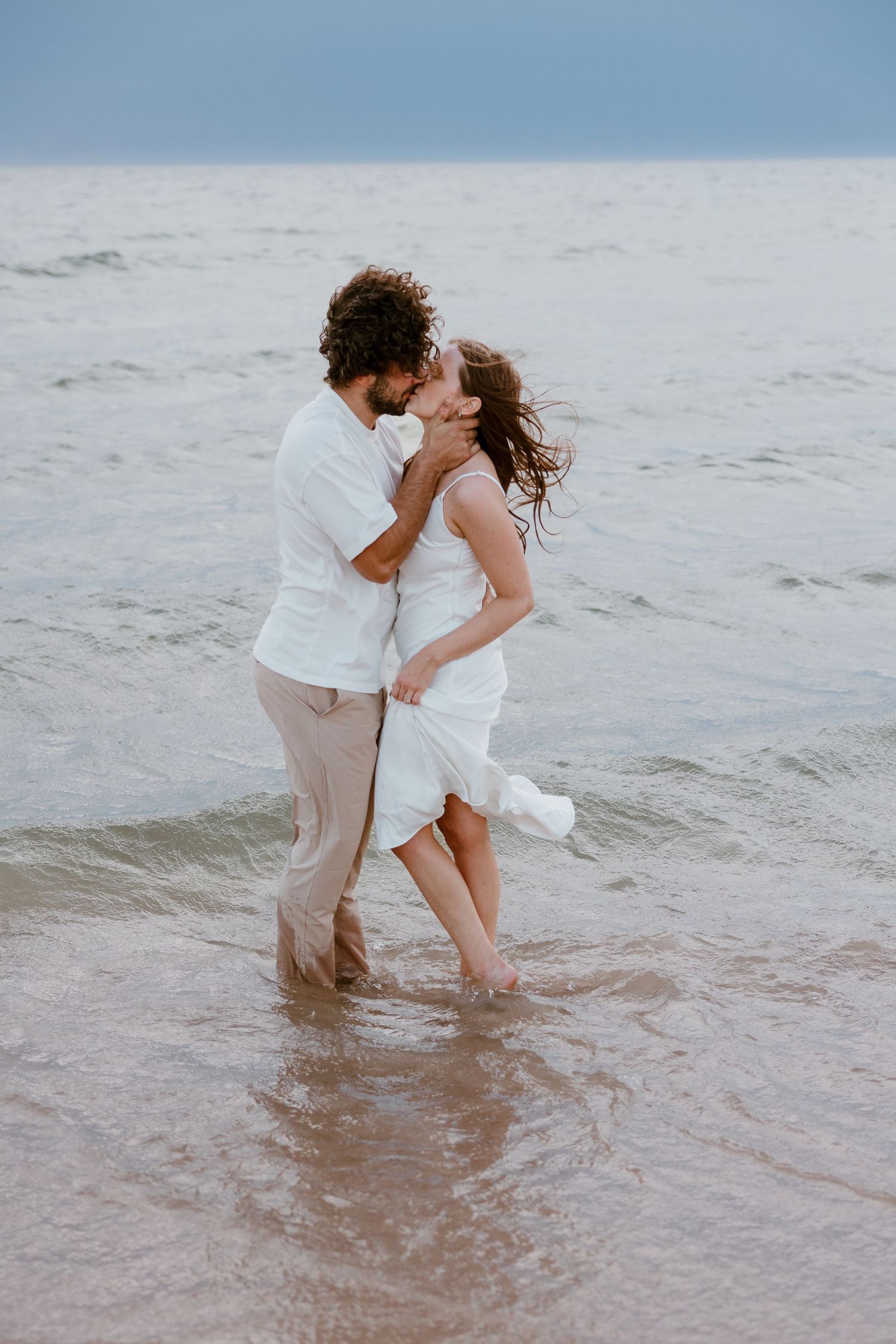 A romantic couple in white clothing embraces and kisses while standing in shallow ocean waves at sunset. Detroit Wedding and Engagement