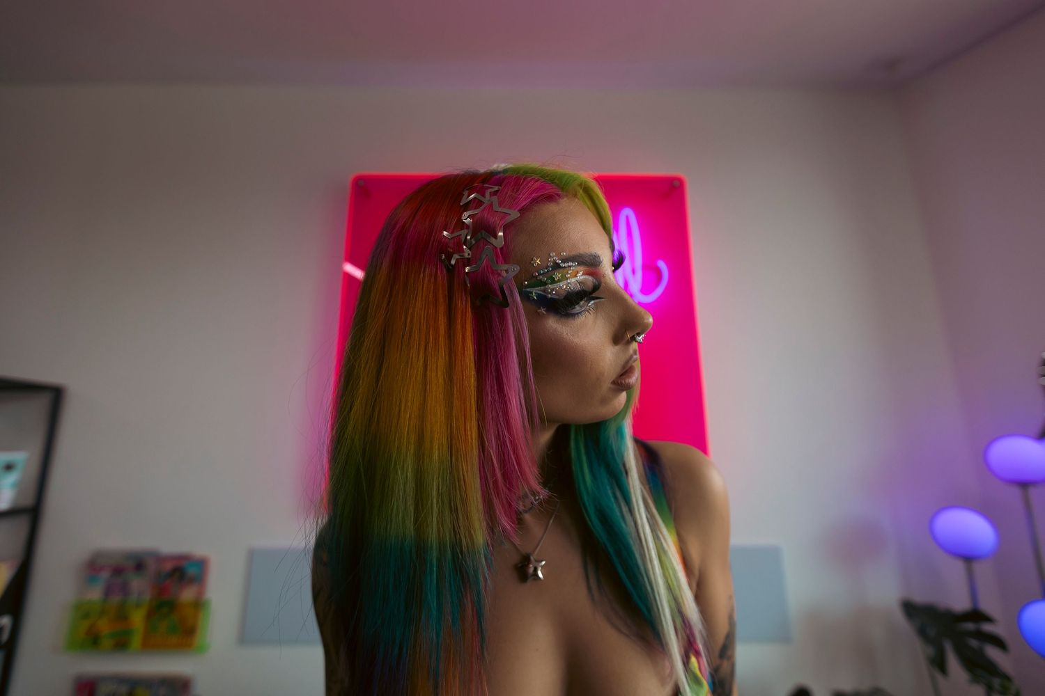 Colorful rainbow hair with face gems glows against pink neon lighting in atmospheric portrait.