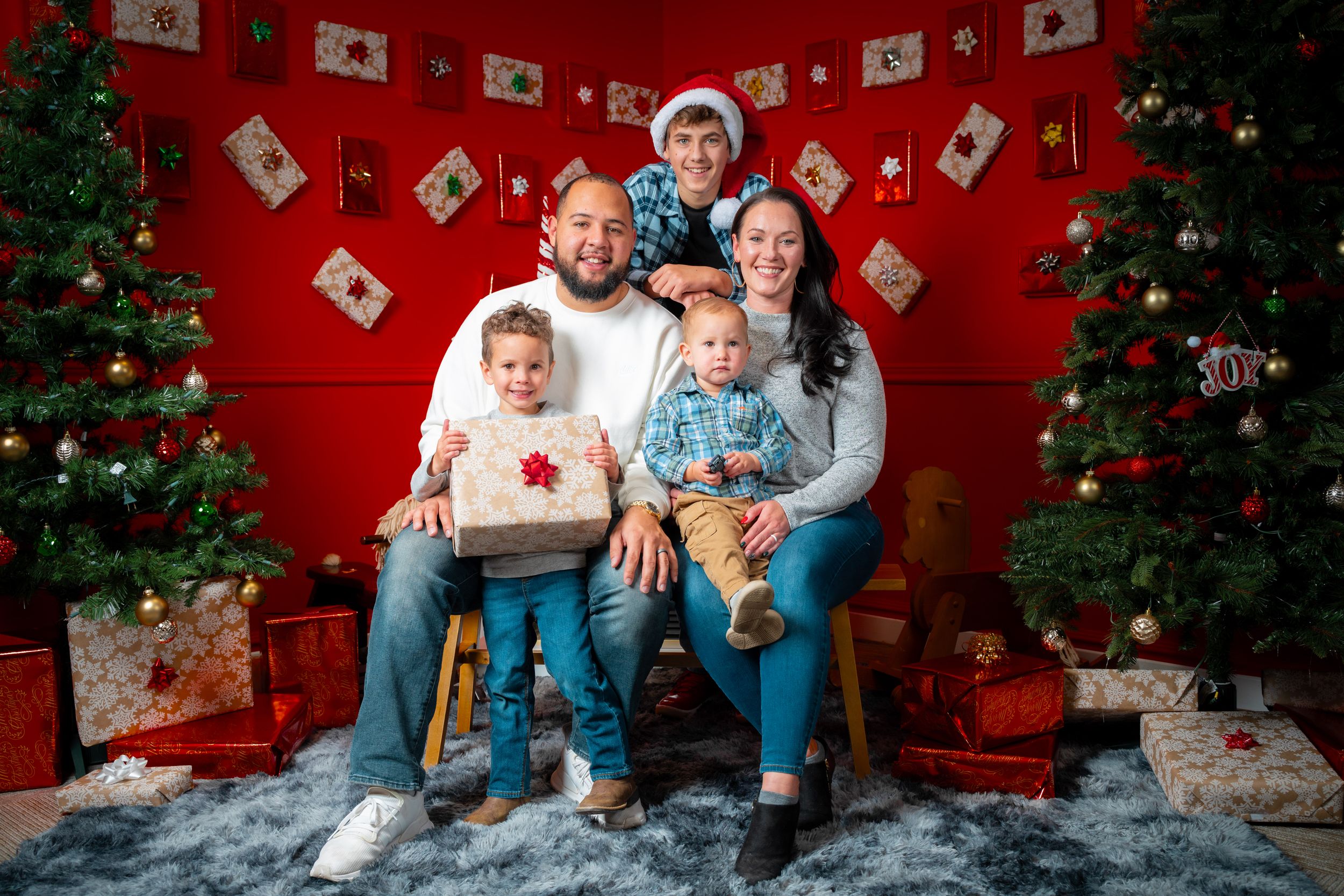 Family poses in matching holiday pajamas with Christmas tree and red wall backdrop for festive portraits.