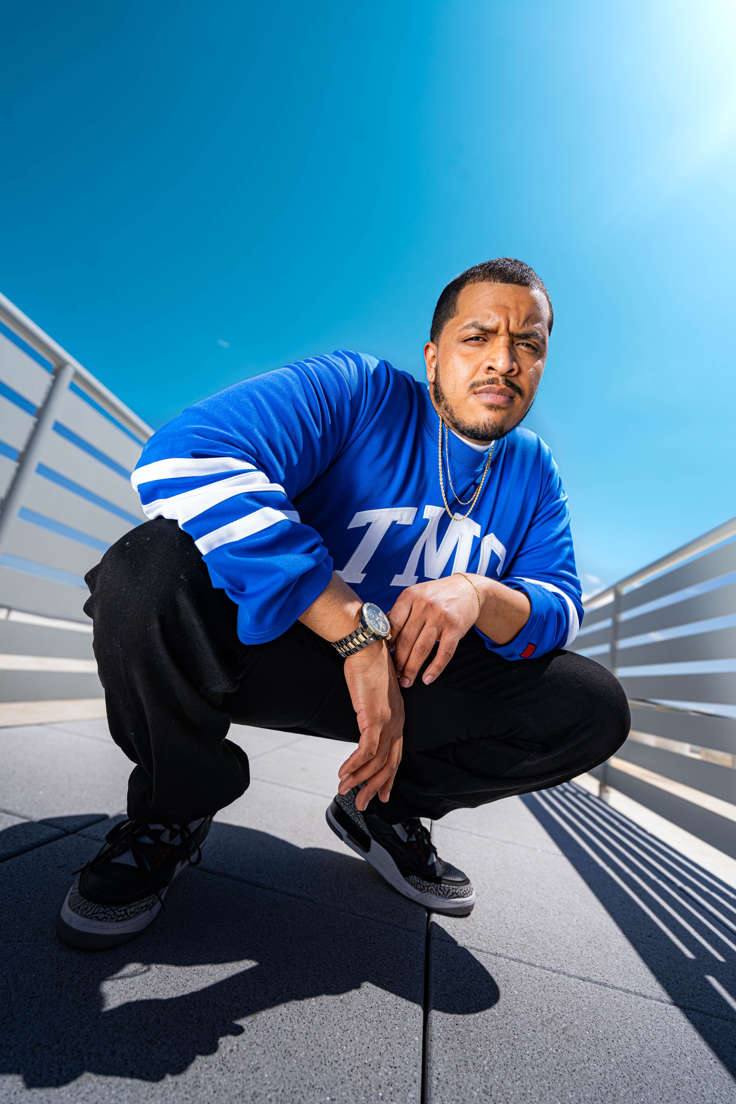 Urban fashion portrait with low angle view of person in blue TMD sweatshirt and black pants crouching on stairs.