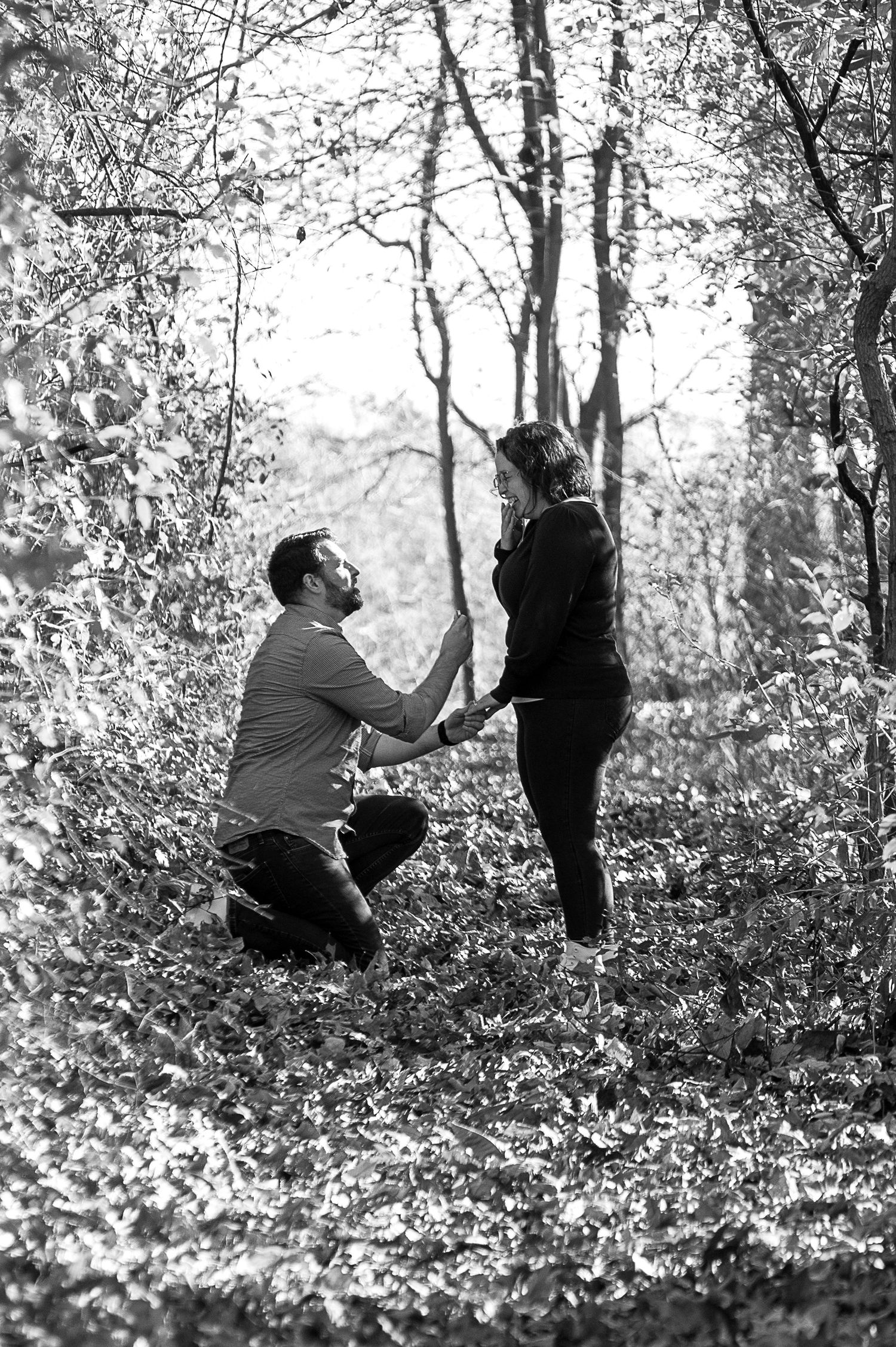 A couple sits on a forest floor surrounded by fallen autumn leaves in black and white.
