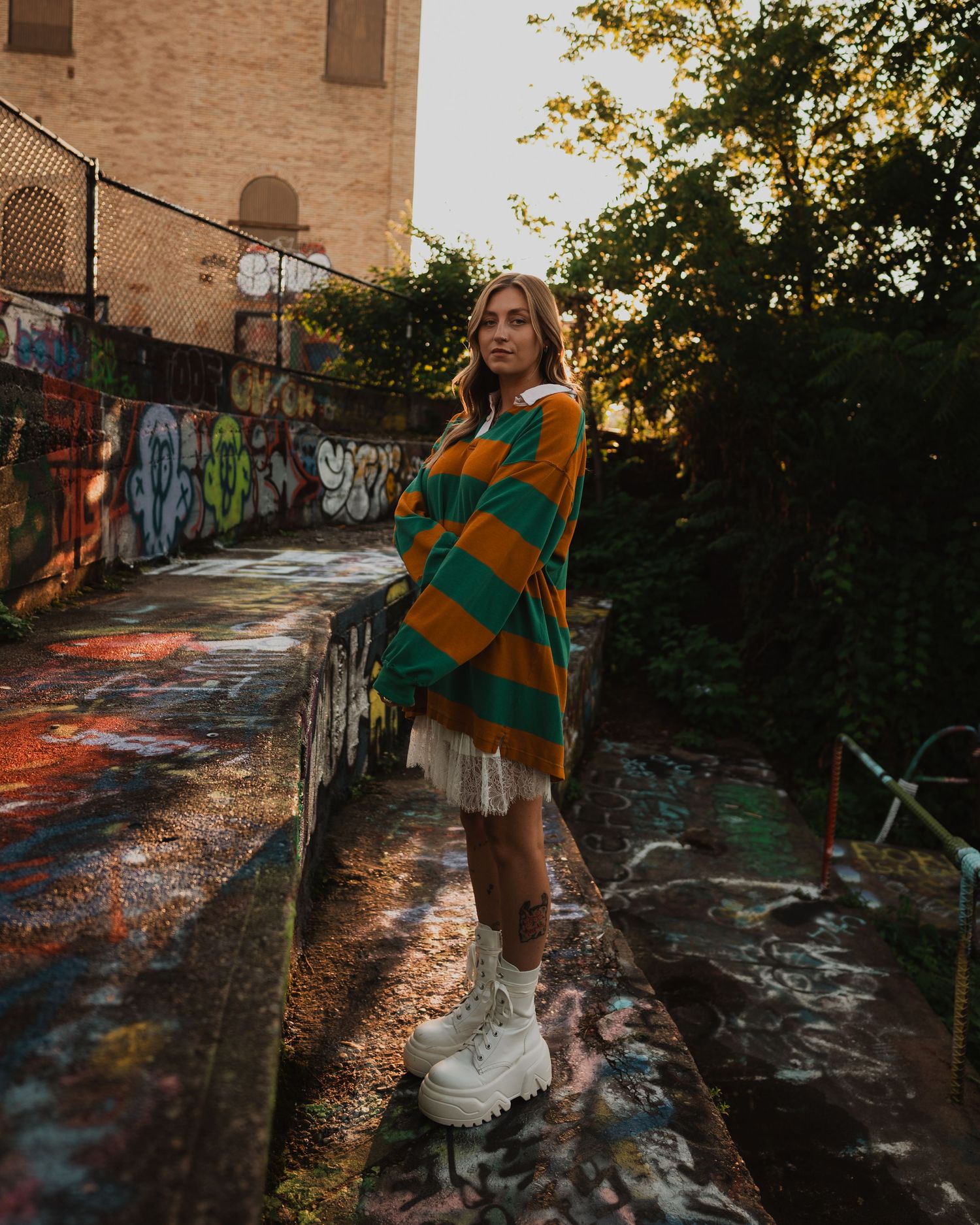 Person in striped green and orange sweater standing on graffiti wall during golden sunset with white sneakers.