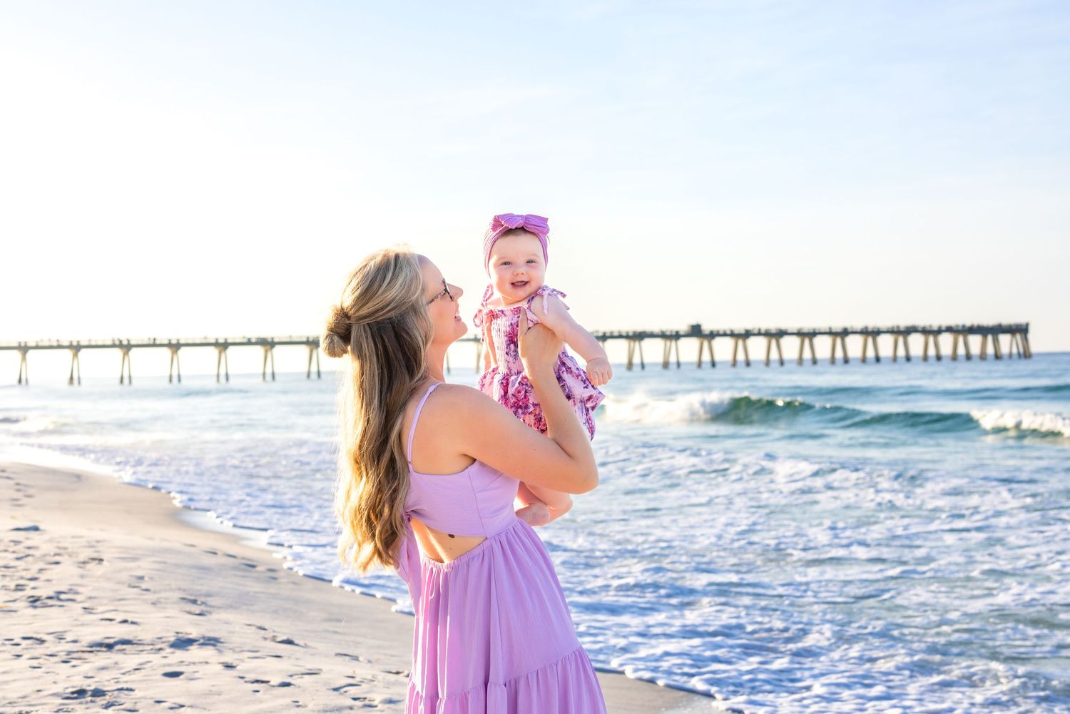 Two people in purple dresses share a loving moment on the beach at sunset with a pier in the background.