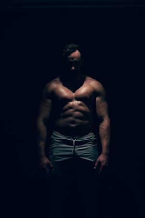 Dramatic low-key portrait of muscular shirtless man standing in dark shadow.