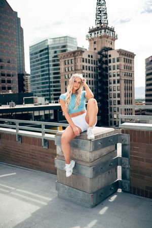 Blonde woman in light blue crop top and white shorts posing on rooftop with Denver city skyline backdrop.