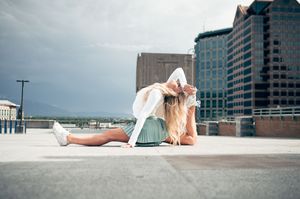 Young blonde woman in white outfit stretching on urban rooftop with city buildings and cloudy sky background.