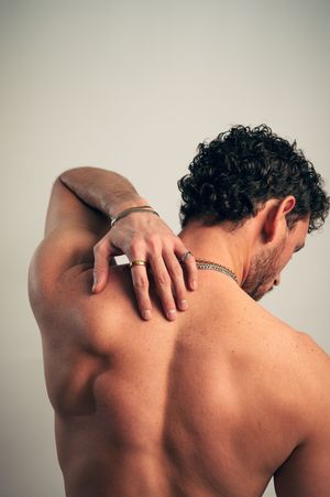 Shirtless curly-haired man touching shoulder, wearing chain necklace and ring.