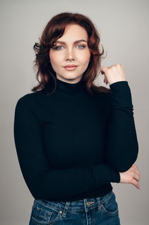 Young woman with wavy auburn hair wearing black turtleneck and jeans posing confidently against gray background.