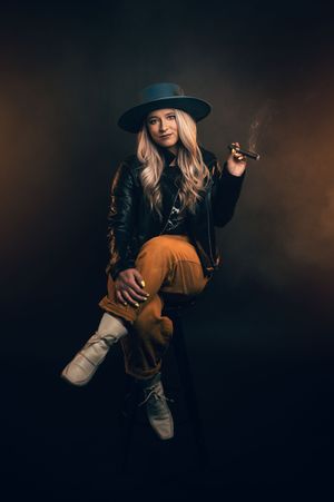 Blonde woman in hat and leather jacket posing with cigar on dark background.
