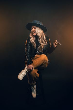 Laughing blonde woman in wide-brim hat holding cigar against dark studio backdrop.