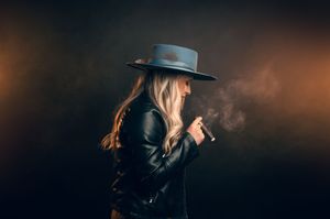Side profile of a blonde woman in a wide-brim hat and leather jacket smoking a cigar against a dark smoky background.
