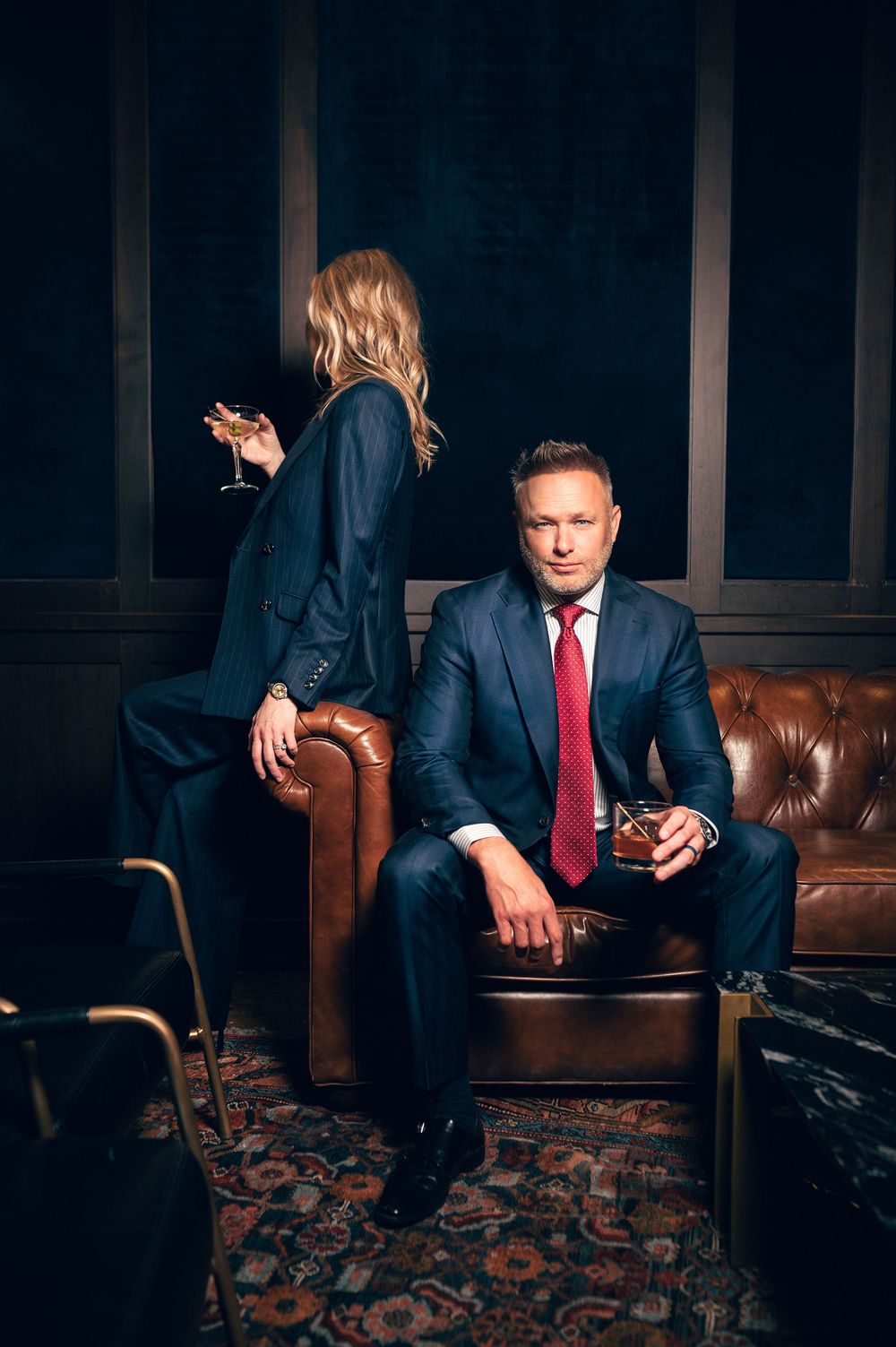 Man in navy suit with red tie seated on leather sofa beside blonde woman in dark elegant room.